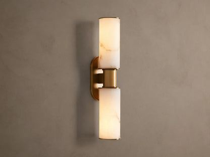 Harlin Double Sconce: Two glowing alabaster cylinder shades with burnished brass trim, mounted vertically on a neutral wall.