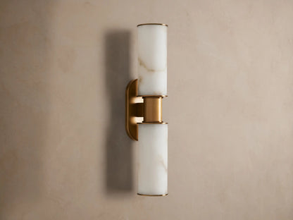 Harlin Double Sconce: Two glowing alabaster cylinder shades with burnished brass trim, mounted vertically on a neutral wall.