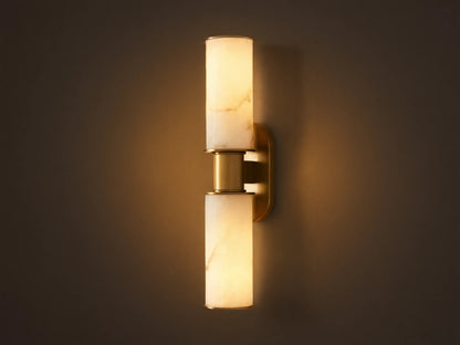 Harlin Double Sconce: Two glowing alabaster cylinder shades with burnished brass trim, mounted vertically on a neutral wall.