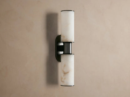 Harlin Double Sconce in Alabaster. Bronze plated fixture with two glowing alabaster cylindrical shades on a taupe wall.