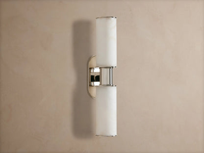 Vertical Harlin Double Sconce in Alabaster with two glowing cylindrical shades and polished nickel on a taupe wall.