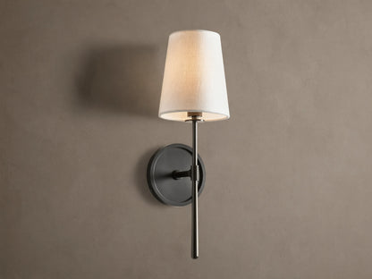 Rhodes Sconce with tapered linen shade on a dark bronze arm and circular backplate, mounted on a light brown wall.