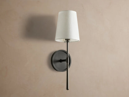 Rhodes Sconce with tapered linen shade on a dark bronze arm and circular backplate, mounted on a light brown wall.