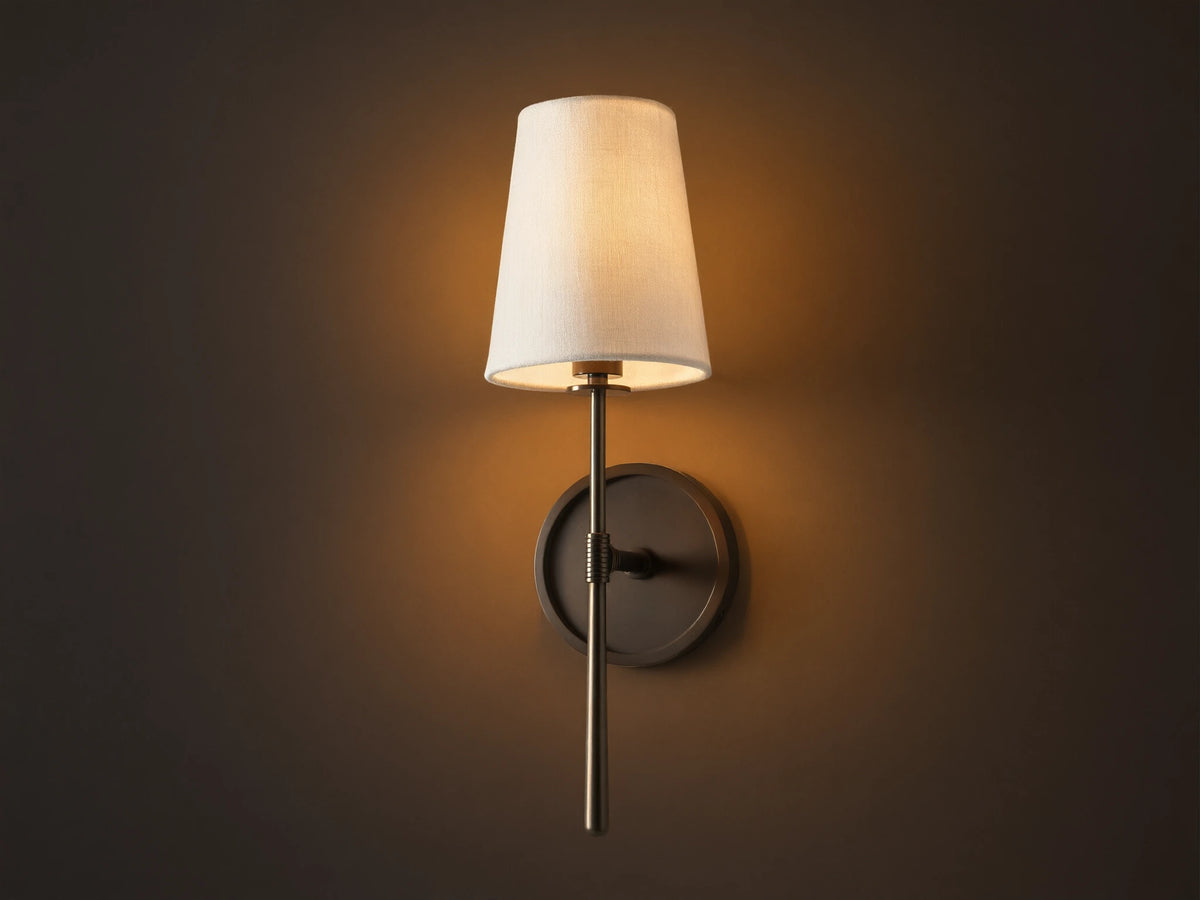 Rhodes Sconce with tapered linen shade on a dark bronze arm and circular backplate, mounted on a light brown wall.