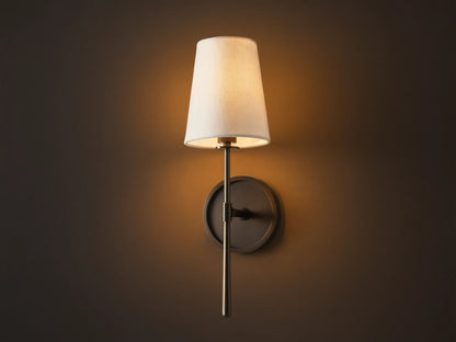Rhodes Sconce with tapered linen shade on a dark bronze arm and circular backplate, mounted on a light brown wall.