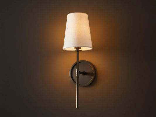 Rhodes Sconce with tapered linen shade on a dark bronze arm and circular backplate, mounted on a light brown wall.