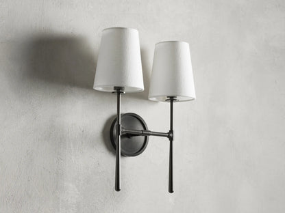 Rhodes Double Sconce with bronze finish, two white tapered linen shades, mounted on a round backplate against a light wall.