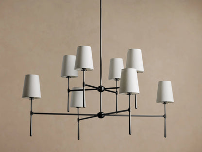 Rhodes Chandelier with 8 light beige tapered linen shades on a dark metal frame, hanging against a tan wall.