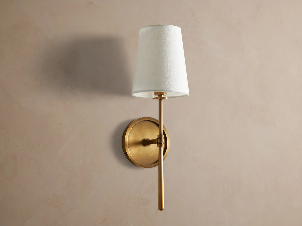 Rhodes Sconce: burnished brass finish wall light, round backplate, and a lit, tapered light linen shade, mounted on a taupe wall.