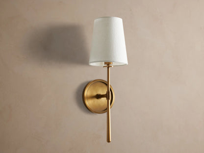Rhodes Sconce: burnished brass finish wall light, round backplate, and a lit, tapered light linen shade, mounted on a taupe wall.