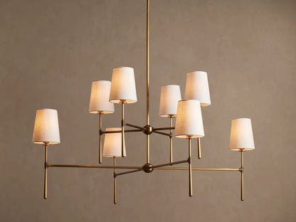 Rhodes Chandelier with burnished brass frame, featuring eight tapered off-white linen shades emitting a warm light.