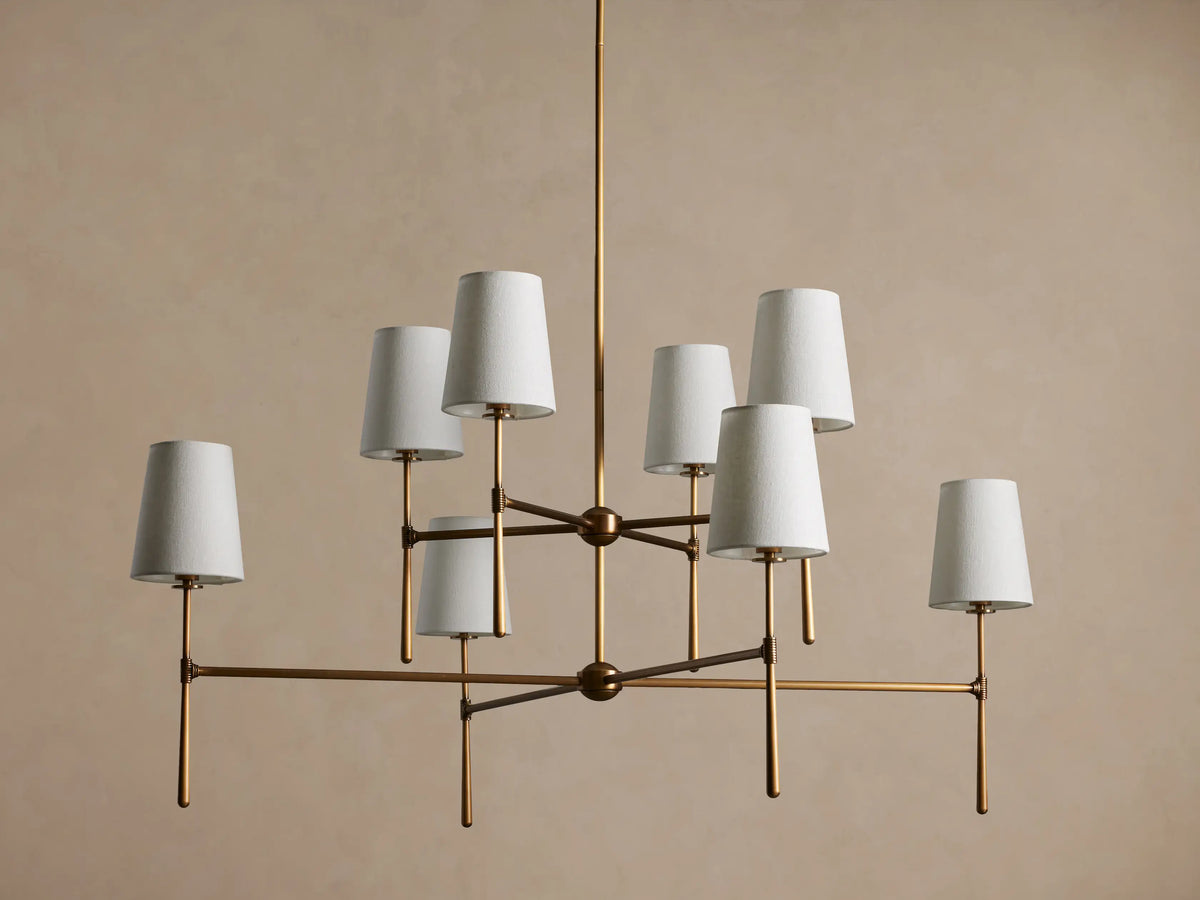 Rhodes Chandelier with burnished brass frame, featuring eight tapered off-white linen shades emitting a warm light.