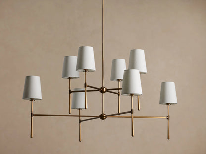 Rhodes Chandelier with burnished brass frame, featuring eight tapered off-white linen shades emitting a warm light.
