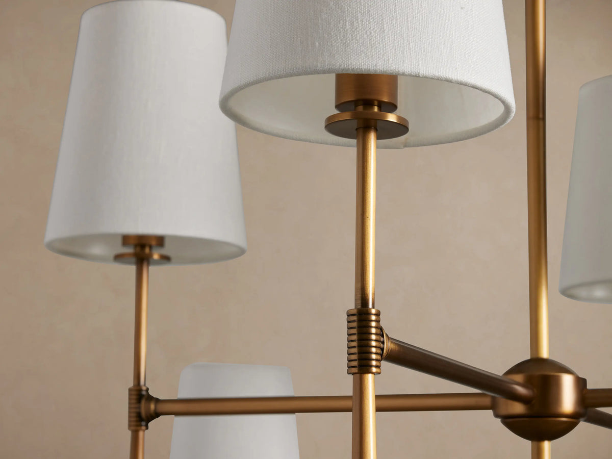 Rhodes Chandelier, burnished brass frame with eight white tapered linen shades, hanging against a light beige wall.