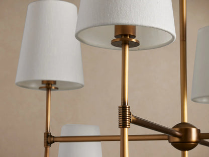 Rhodes Chandelier, burnished brass frame with eight white tapered linen shades, hanging against a light beige wall.