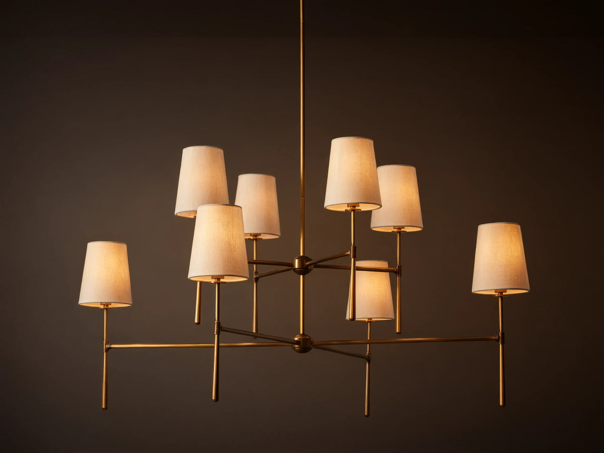 Rhodes Chandelier with burnished brass frame, featuring eight tapered off-white linen shades emitting a warm light.