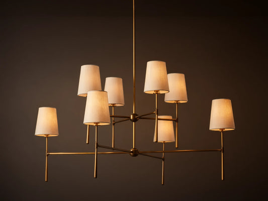 Rhodes Chandelier with burnished brass frame, featuring eight tapered off-white linen shades emitting a warm light.