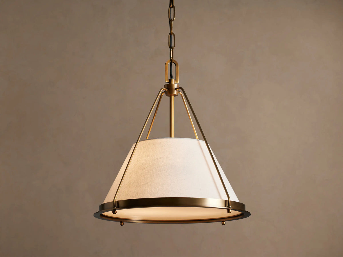 Shelton Pendant: burnished brass metal frame & chain, holds glowing tapered off-white linen shade, hanging against beige wall.