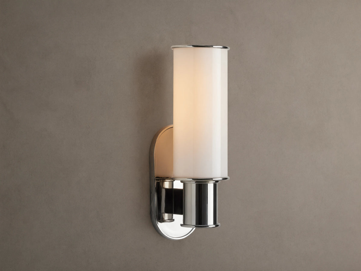 Harlin Sconce in Milk. Polished nickel wall sconce with a lit, vertically oriented milk glass cylinder on a beige wall.