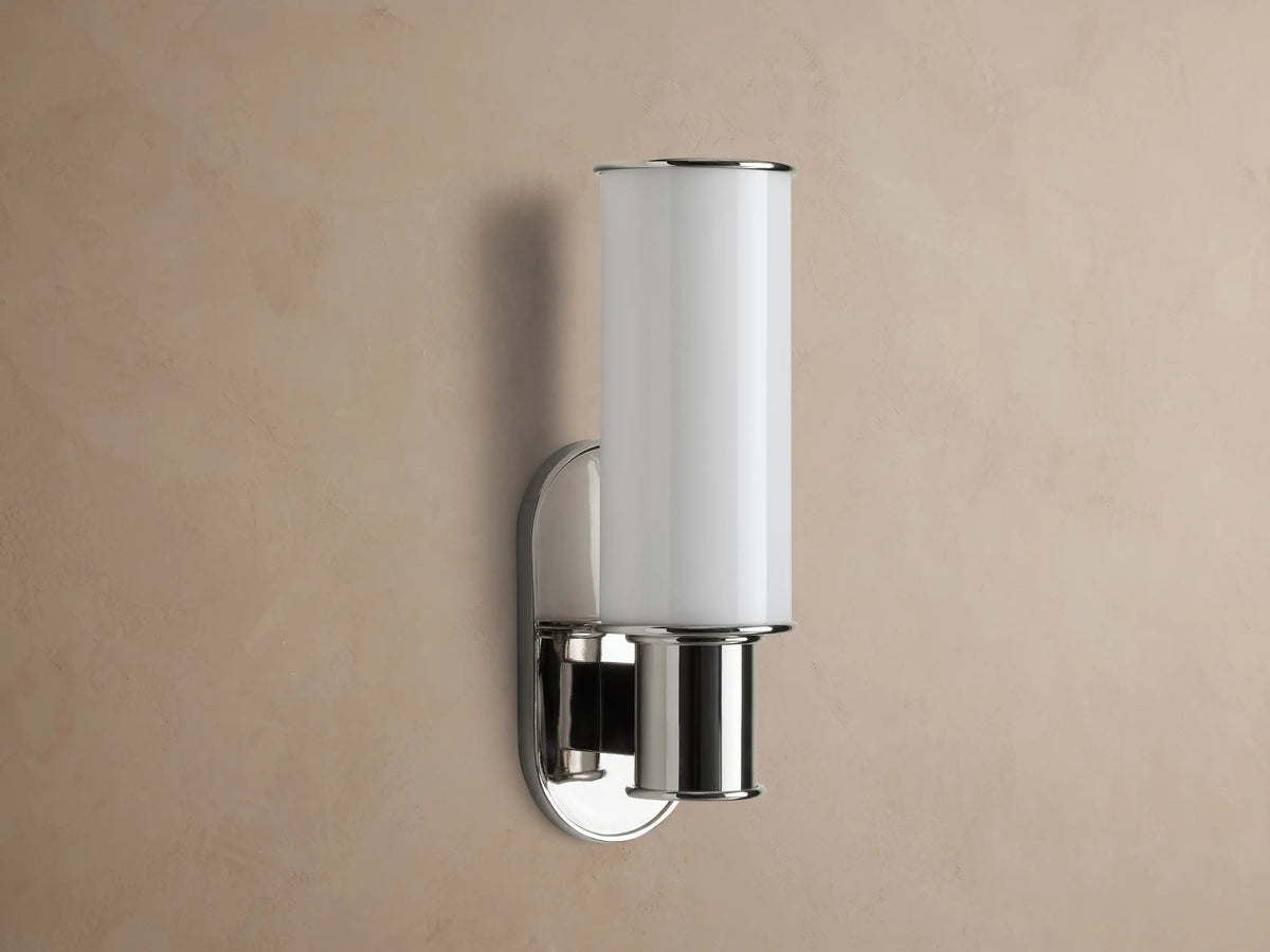 Harlin Sconce in Milk. Polished nickel wall sconce with a lit, vertically oriented milk glass cylinder on a beige wall.