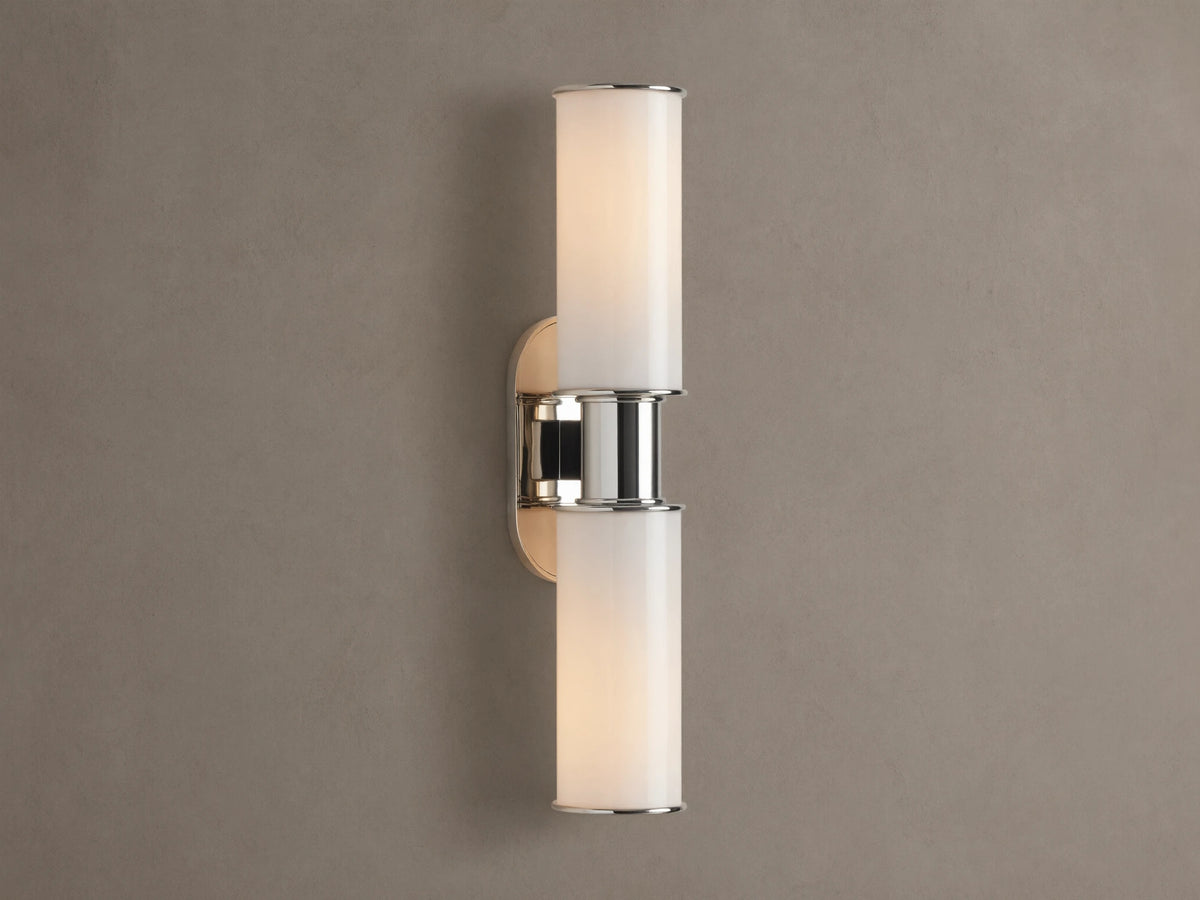 Harlin Double Sconce with two glowing vertical milk glass shades and polished nickel accents, mounted on a textured brown wall.
