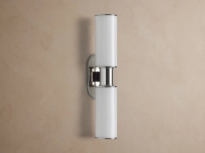 Harlin Double Sconce with two glowing vertical milk glass shades and polished nickel accents, mounted on a textured brown wall.