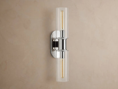 Vertical Harlin Double Sconce with polished nickel finish and two clear glass cloches revealing illuminated Edison bulbs on a brown wall.