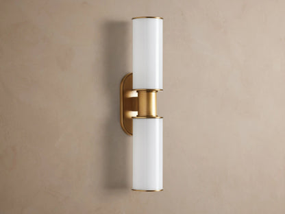Harlin Double Sconce mounted vertically on a wall. It has two glowing milk glass cylinders separated by a burnished brass stem & oval backplate.