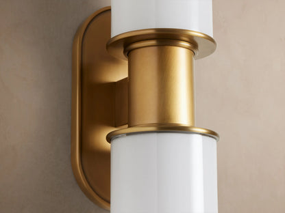 Harlin Double Sconce in Milk with two vertical white cylindrical glass shades and burnished brass details on an oval backplate.