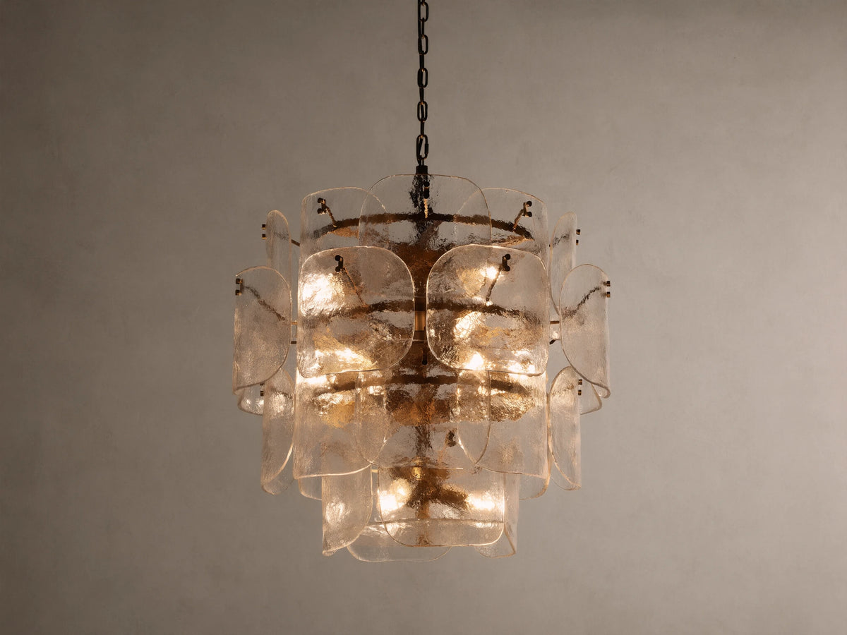 Blaire Round Chandelier with layered, curved clear glass panels creating a textured effect. Lit with warm light, hung by a brass-finished chain.