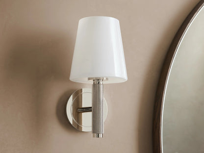 Monroe Sconce in polished nickel finish with a lit white glass shade, mounted on a beige wall beside a mirror.