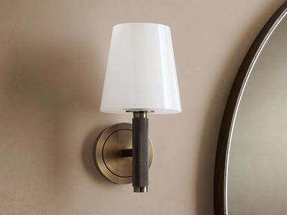 Monroe Sconce with antique brass finish, round wall mount, and soft glowing milk glass shade, shown beside mirror.