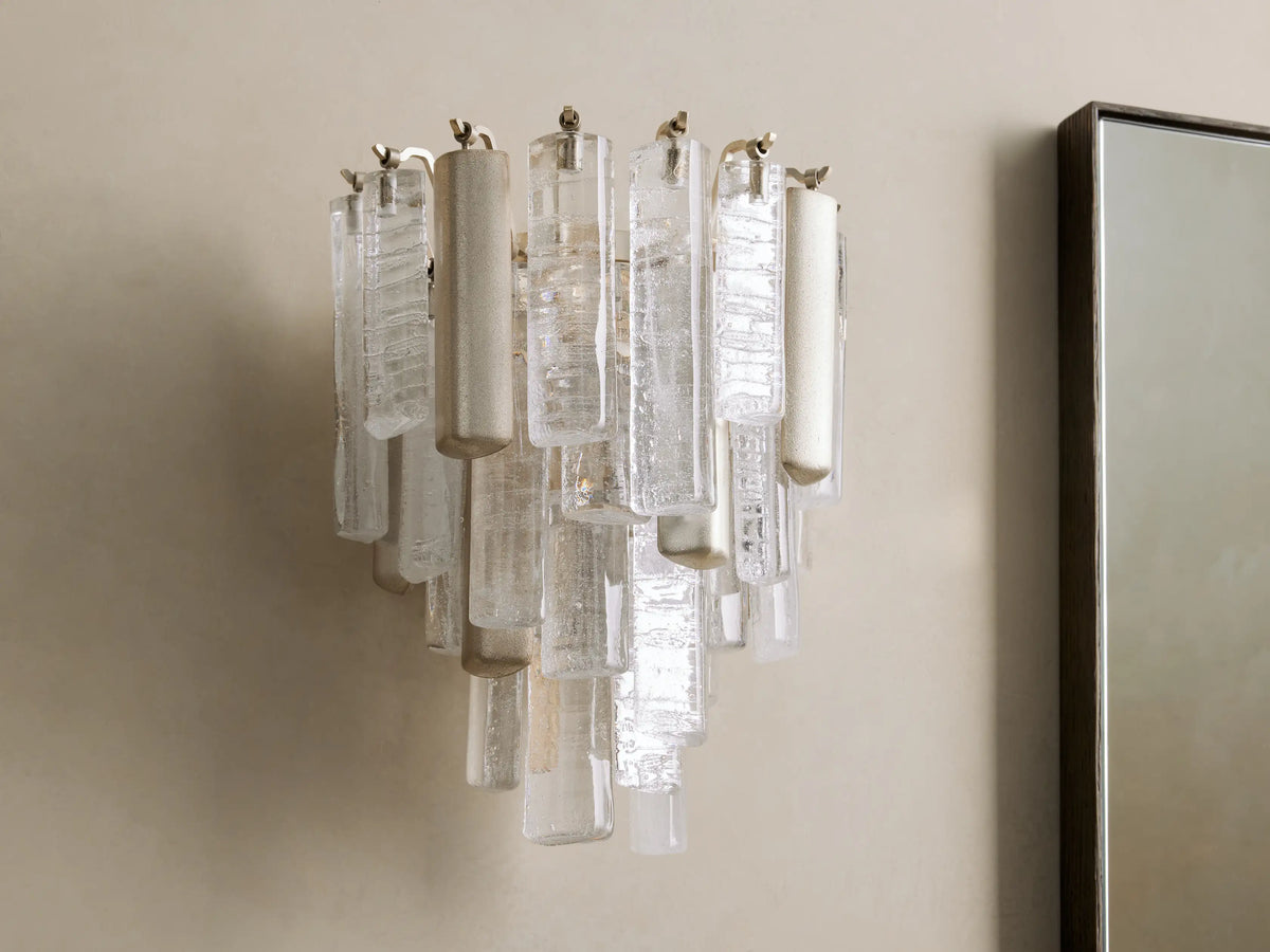 Neve Sconce: Wall-mounted light with clear textured glass and champagne-finished aluminum elements, glowing warmly.