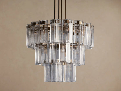 Delsie Round Chandelier, multi-tiered with clear ribbed glass rods and burnished brass frame, hanging against a brown wall.