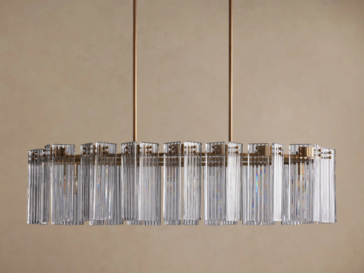 Delsie Oval Chandelier with clear ribbed glass rods and a burnished brass frame, hanging against a light brown wall.