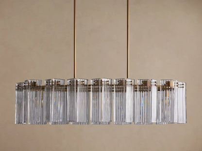 Delsie Oval Chandelier with clear ribbed glass rods and a burnished brass frame, hanging against a light brown wall.