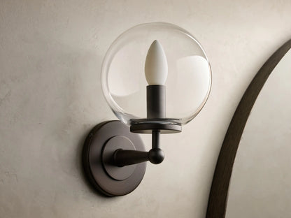Close-up of the antique bronze Fulton Sconce with a clear glass globe, mounted on a textured wall.