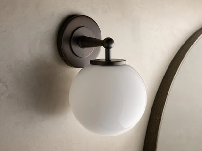 Close-up of the antique bronze Fulton Sconce with a glowing milk glass globe attached to a round backplate.