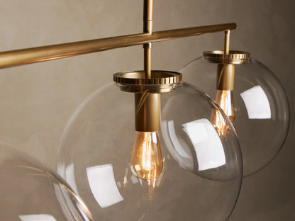 Front view of a linear pendant light featuring three clear glass globe shades, brass hardware, and exposed filament bulbs.