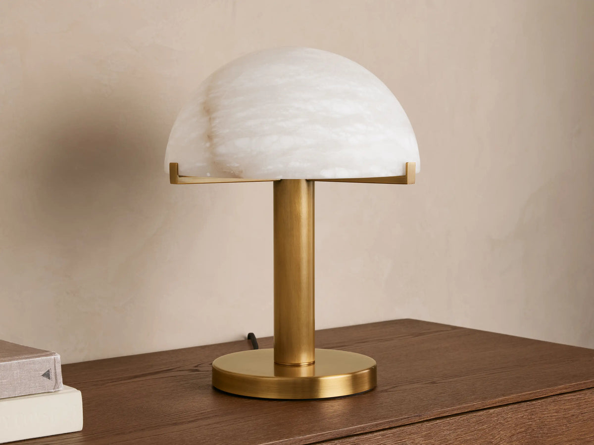 Emerie Table Lamp with a dome-shaped alabaster shade on a brass base, placed on a wooden surface against a neutral wall.