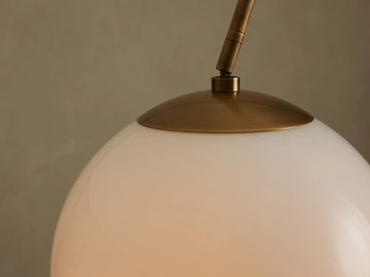 Close-up of Christie Floor Lamp's glowing milk glass shade with a brass cap and arm extending up-left, against a neutral wall.
