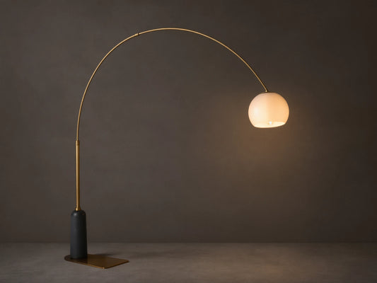 Christie Floor Lamp: Arched brass-finished lamp with glowing milk glass shade, dark stone base on wood floor against beige wall.