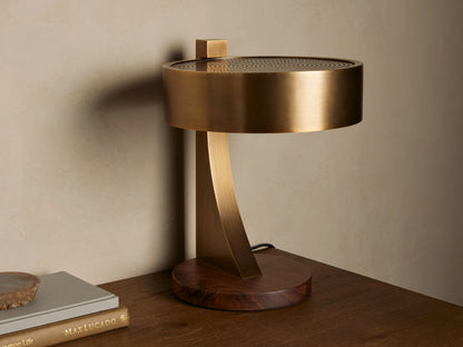 Alastair Table Lamp with a burnished brass cylindrical shade, perforated top, curved arm, and oiled walnut base, emitting warm light on a wooden table.