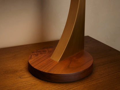 Alastair Table Lamp on a wooden table. Dark brown walnut base, curved burnished brass stem, circular shade with perforated top.