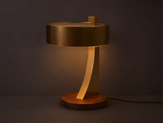 Alastair Table Lamp with a burnished brass cylindrical shade, perforated top, curved arm, and oiled walnut base, emitting warm light on a wooden table.
