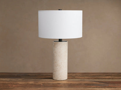 Brixton Table Lamp, front view. Cylindrical light beige marble base, dark metal stem, white fabric drum shade, on wood.