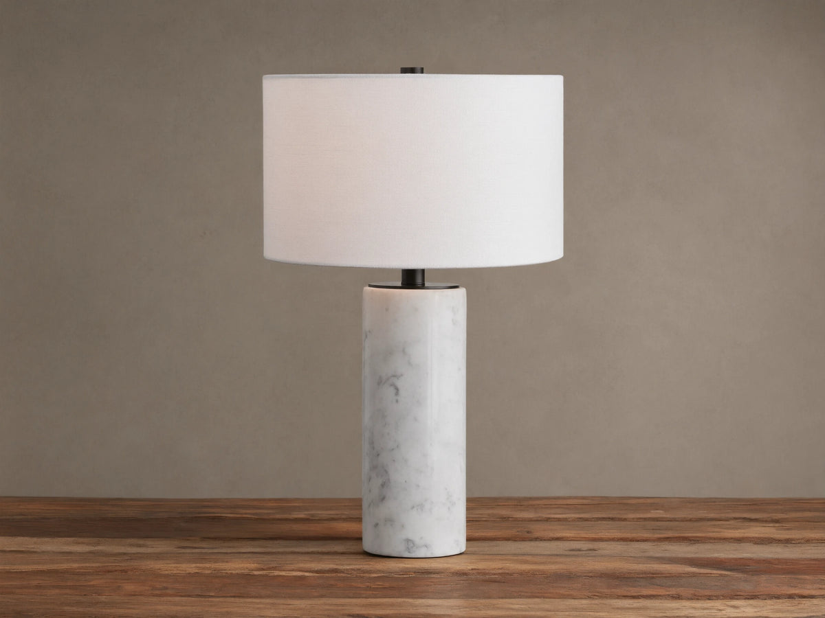 A Brixton Table Lamp featuring a cylindrical white marble base, black stem, and white fabric shade on a wooden table.