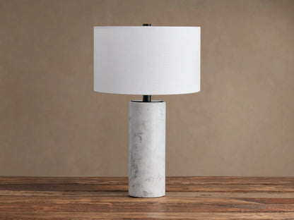 A Brixton Table Lamp featuring a cylindrical white marble base, black stem, and white fabric shade on a wooden table.