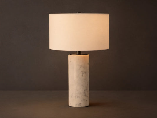 A Brixton Table Lamp featuring a cylindrical white marble base, black stem, and white fabric shade on a wooden table.