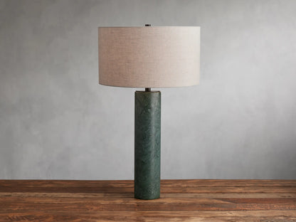 Brixton Table Lamp with a cylindrical green marble base and a large cream fabric lampshade, placed on a wooden surface against a gray wall.